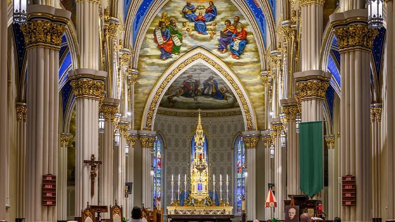 The Basilica of the Sacred Heart has ornate white columns with gold and blue details on either side of the nave. The tall, arched ceilings are covered in elaborate murals depicting heavenly scenes. A Catholic alter with three candles on each side of a tall golden tabernacle tower is the central focus of the image.
